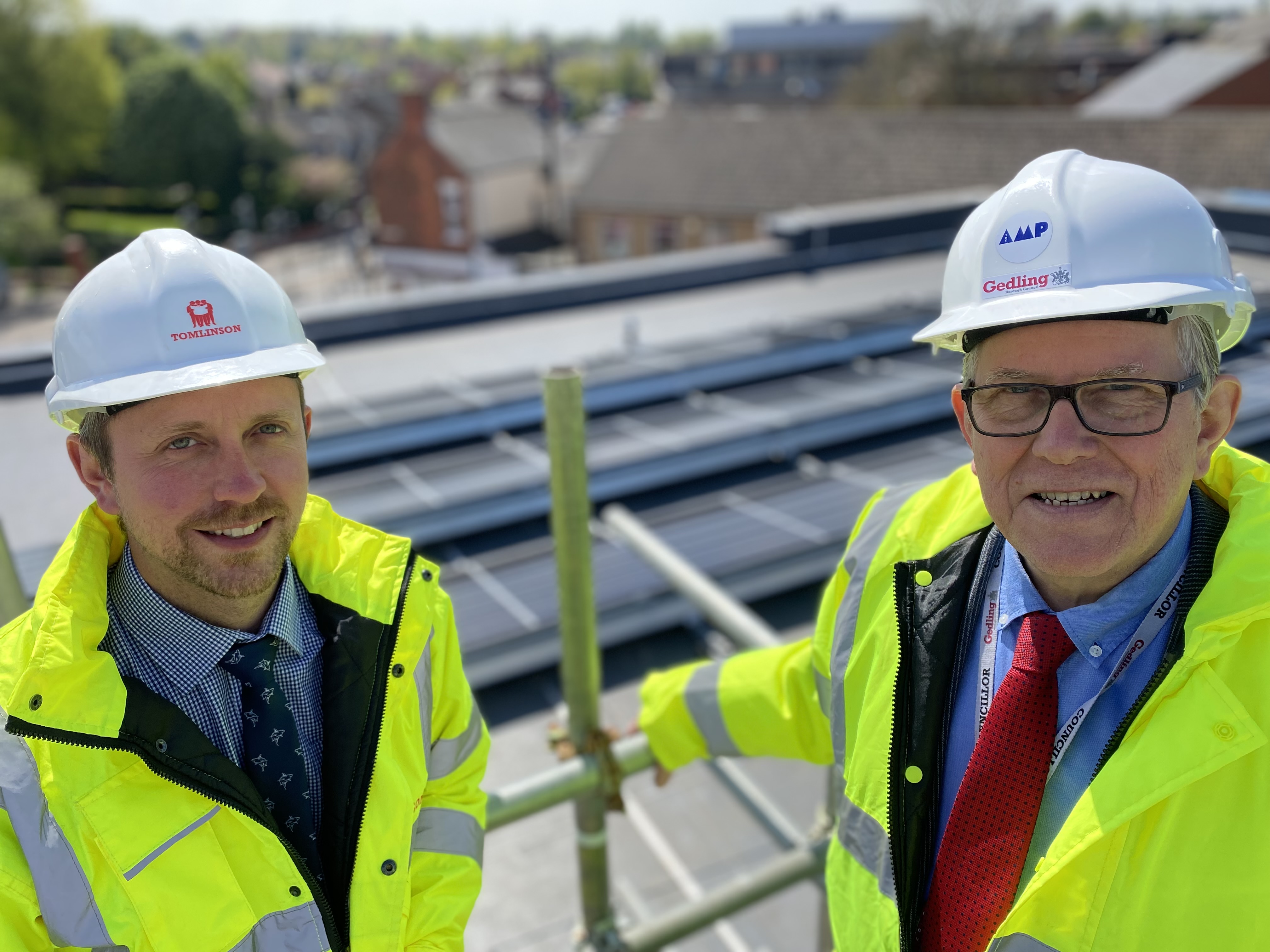 Two men wearing hard hats and high visibility jackets standing on the roof of the building with the solar panels in the background.